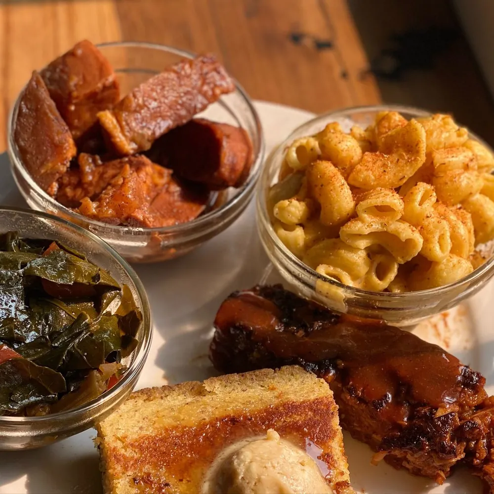 A platter of yams, mac and cheeze and collard greens in glass bowls, with cornbread, maple butter and BBQ ribs